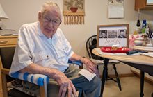 Lester Donley, DO, (COM 1952) sits at a kitchen table smiling next to a family photo. 